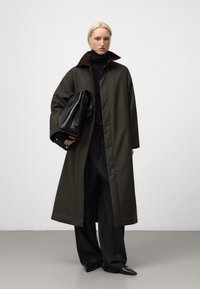 Cappotto lungo verde oliva scuro con colletto bordeaux, abbinato a un dolcevita nero e pantaloni larghi neri. Tiene una borsa in pelle nera.