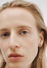 Pendientes colgantes de oro que presentan una piedra preciosa morada como piedra de fondo y un diseño esbelto y alargado con un acento curvado en la parte inferior.