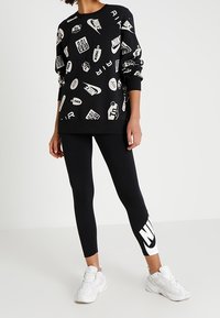 Černá oversized mikina s bílými logy a grafikou Nike, zkombinovaná s černými legínami s velkým bílým logem Nike. Bílé tenisky.