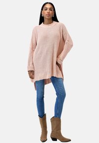 Heller rosa, übergroßer Strickpullover mit einem gerippten Ausschnitt, langen Ärmeln und seitlichen Schlitzen, kombiniert mit taillierten blauen Jeans und braunen Stiefeletten.