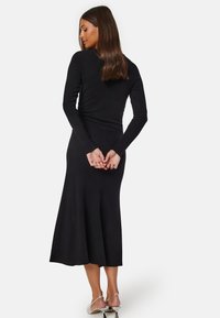 Zwarte geribbelde jurk met lange mouwen, nauwsluitend lijfje en uitlopende midi-lengte rok. Glad textuur, minimale details en onzichtbare rits sluiting.