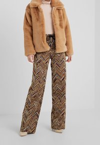 Braune Kunstfelljacke mit weicher Textur; gemusterte weit geschnittene Hose mit Zickzacklinien in den Farben Gold, Braun und Rost.