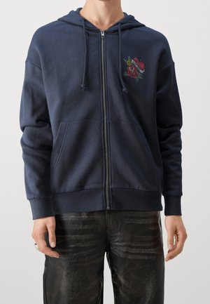 Navy zip-up hoodie made of soft fabric, featuring front pockets and an embroidered rose design on the left chest. Textured cuffs with drawstrings.