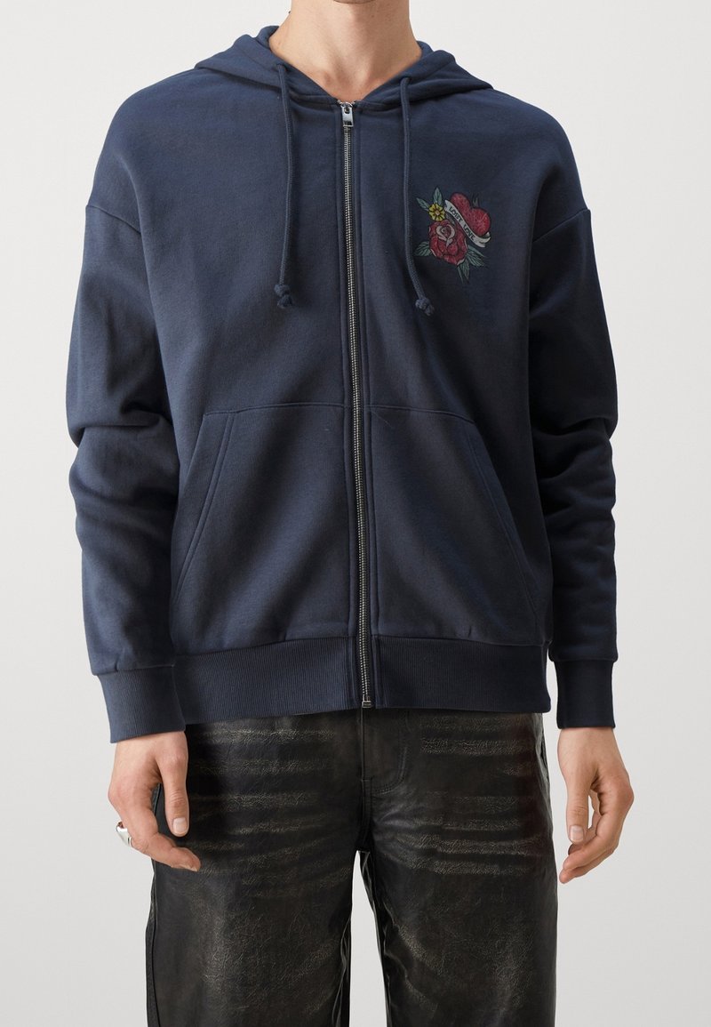 Felpa con zip color navy realizzata in morbido tessuto, dotata di tasche frontali e di un design di rosa ricamato sul lato sinistro del petto. Polsini strutturati con cordini.
