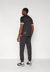 Camiseta preta com detalhes dourados e mangas curtas, combinada com calças de treino cinza escuro com uma risca lateral estampada. Tecido texturizado.