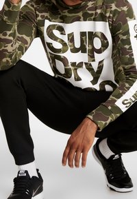 Sudadera verde de camuflaje con logo blanco de "Sup Dry"; joggers negros ajustados; zapatos negros de Nike; calcetines blancos visibles; estilo atlético casual.
