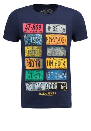 Mężczyzna w granatowej koszulce z nadrukiem kolorowych tablic rejestracyjnych, podwiniętych dżinsach, szarych tenisówkach, trzymający brązowy plecak przy boku.