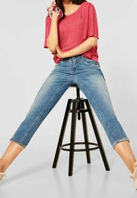 Niebieskie jeansy z denimu o skróconej długości i podwiniętych mankietach, z efektem wyblakłego prania. Zestawione z czerwonym topem z krótkim rękawem.