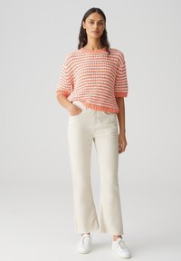 Pull en tricot avec des rayures orange et blanches, coupe décontractée ; associé à un pantalon évasé de couleur claire et des baskets blanches.