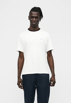 Joven de piel oscura y rastas, vistiendo una camiseta blanca de manga corta y pantalones oscuros, de pie contra un fondo liso.