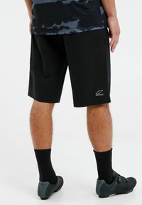 Mustat urheilushortsit, joissa on sileä pinta, polvipituinen muotoilu, ja alareunassa on pieni valkoinen logo. Vaatteena mustat sukat ja kengät.