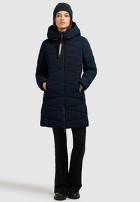 Abrigo puffer azul marino con diseño acolchado, capucha, cremallera frontal y dos bolsillos laterales. Combinado con pantalones negros y un gorro de punto oscuro.