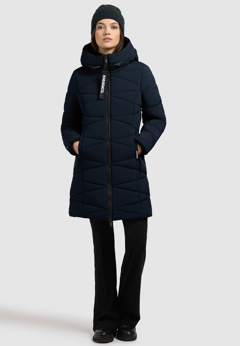 Abrigo puffer azul marino con diseño acolchado, capucha, cremallera frontal y dos bolsillos laterales. Combinado con pantalones negros y un gorro de punto oscuro.