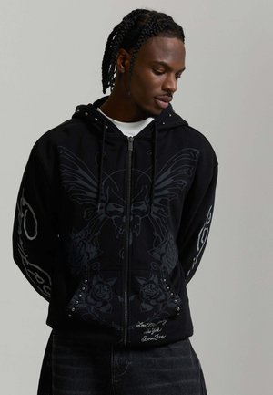 Junger Mann mit geflochtenem Haar, der eine schwarze Zip-Hoodie mit einem Schädel- und Flügeldesign mit floralen und textlichen Details trägt.