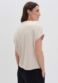 Donna con capelli castani lisci che indossa un top beige a maniche corte e ampio e pantaloni neri, in piedi di profilo con le mani in tasca su uno sfondo semplice.