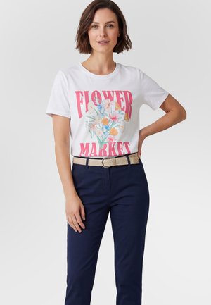 Kvinde med kort brunt hår iført en hvid T-shirt med farverige blomster og teksten "FLOWER MARKET", navybukser og et beige vævet bælte.