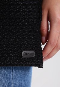 Hand hält den Rand eines schwarzen, strukturierten Strickpullovers mit dem Replay-Logo, über blauen Jeans.