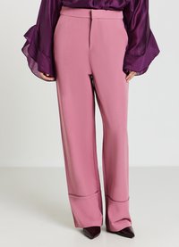 Frau in hochgeschnittenen, weiten, pinken Hosen mit einer Nahtdetails nahe dem Saum, kombiniert mit einer lila Satinbluse und spitzen Schuhen.