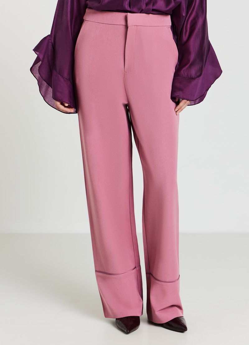 Frau in hochgeschnittenen, weiten, pinken Hosen mit einer Nahtdetails nahe dem Saum, kombiniert mit einer lila Satinbluse und spitzen Schuhen.