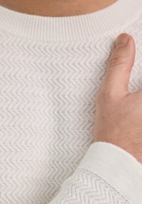 Texturiertes weißes Sweatshirt mit einem Zickzackmuster, das über einen gerippten Halsausschnitt und einen Rippabschluss verfügt. Eine Hand ruht auf dem Schulterbereich und zeigt die Details des Kleidungsstücks.