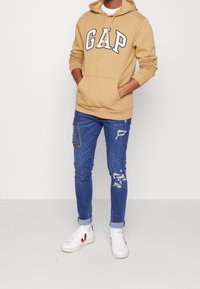 Sweat à capuche beige avec "GAP" en lettres majuscules, accompagné d'un jean bleu slim-fit avec des zones déchirées et une poche patch, et des baskets blanches.