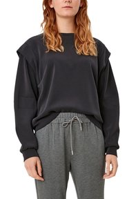 Sudadera negra con hombros caídos y bolsillo lateral, combinada con pantalones de chándal grises con cordón. Tejido suave con un ajuste casual.