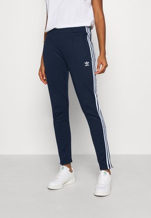 Personne portant un pantalon de survêtement Adidas bleu marine avec des bandes blanches sur les côtés et des baskets blanches, debout devant un fond uni.