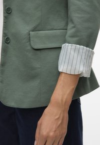 Personne portant un blazer vert avec des poignets de chemise rayés blancs retroussés, montrant le rabat de la poche et une partie du pantalon bleu foncé.