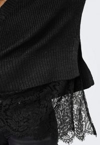 Pull en maille côtelée noire superposé sur un tissu en dentelle florale noire avec des bords festonnés, vu de la taille vers le bas sur une personne.