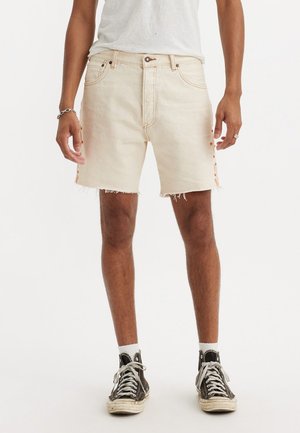 Shorts de mezclilla beige con dobladillo deshilachado y acentos de botones laterales. Llevados con zapatillas altas grises y calcetines blancos. Diseño simple.