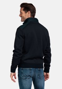 Marineblauwe sweatshirt met opstaande kraag, groene accenten, geribde manchetten en zoom. Zacht materiaal, casual pasvorm. Gekleed met blauwe jeans.