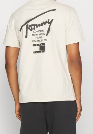 Roomkleurig katoenen T-shirt met een zwarte grafische print op de achterkant met "Tommy" en stadsnamen, samen met "TOMMY JEANS" in een kader.