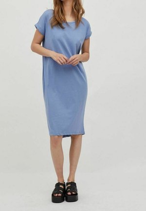 Kvinde iført en lyseblå knælængde kjole med korte ærmer og sorte plateau-sandaler, stående mod en enkel lysegrå baggrund.