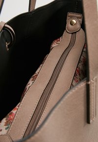 Beige laukku, jossa on teksturoitu ulkopinta ja musta sisus; sisältää kukkakuosisen vuorin, vetoketjullisen osaston ja kultasävyisiä yksityiskohtia.