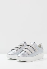 Metallische zilveren sneakers met drie klittenbandstraps. Voorzien van gestructureerde panelen en een witte rubberen zool. Minimalistisch ontwerp, geen veters.