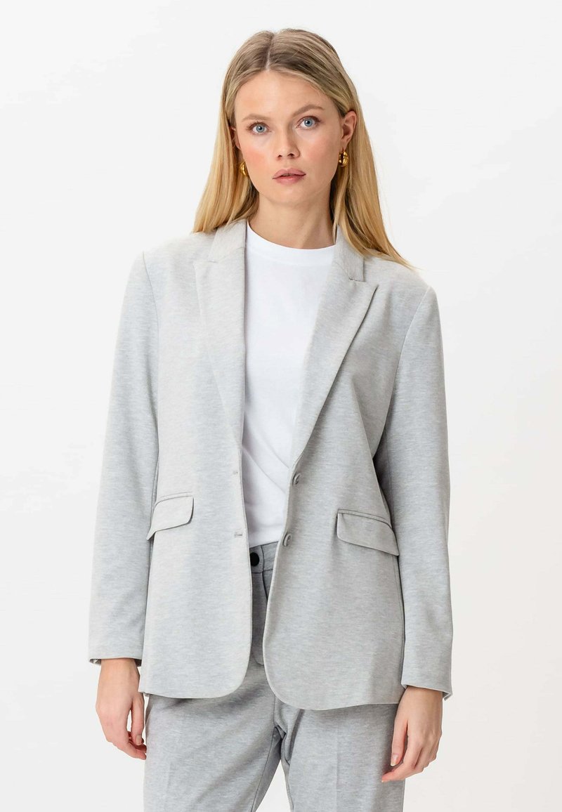 Femme blonde portant des boucles d'oreilles en or, un blazer gris clair avec des poches à rabat sur une chemise blanche et un pantalon gris assorti, se tenant devant un fond blanc.