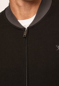 Zwarte jas met rits en een grijze geribbelde kraag. Heeft een gladde textuur en een klein logo op de borst vlakbij de rits.