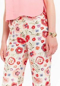 Pantalons à motif floral avec un fond crème, ornés de fleurs rouges, roses et bleues. Fermeture à cinq boutons à la taille, tissu léger.