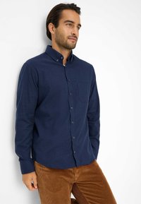 Uomo che indossa una camicia azzurra con bottoni e pantaloni di corduroy marroni, in piedi contro un muro bianco, che guarda leggermente a destra.