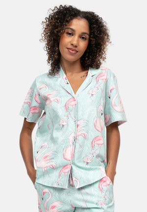 Frau mit lockigem Haar, die ein hellblaues, kurzärmliges Hemd mit Knöpfen und eine Hose mit pinkem Flamingo-Print trägt, steht mit den Händen in den Taschen.