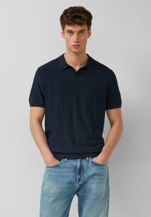 Junger Mann mit kurzen braunen Haaren, der ein dunkelblaues kurzärmeliges Poloshirt und hellblaue Jeans trägt, die Hände in den Taschen.