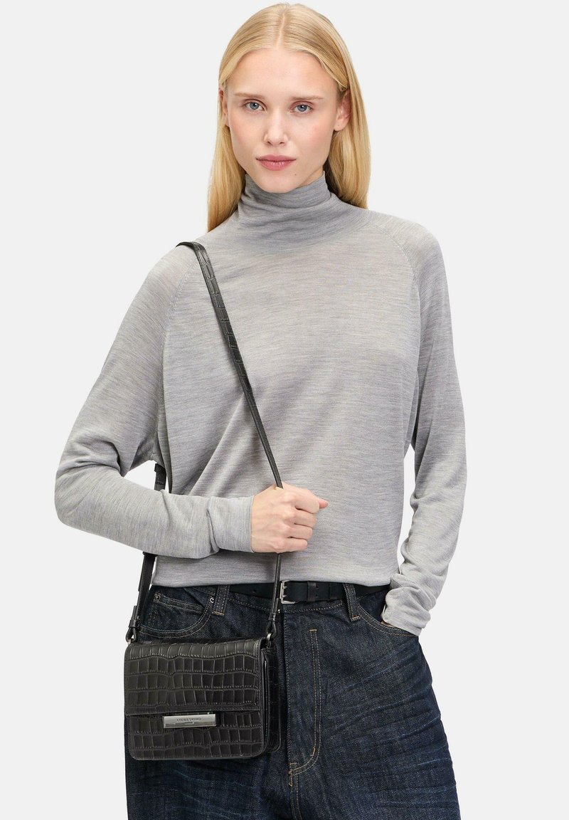 Schwarze, aus Krokodilleder geprägte Umhängetasche mit silberfarbener Hardware. Das Model trägt ein graues Longsleeve mit hohem Kragen und dunklen Jeans.