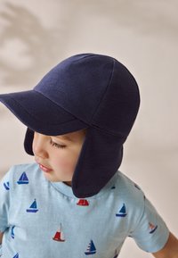 Gorra de algodón azul marino con solapas y visera curva, usada por un niño. La camiseta debajo presenta un fondo azul claro con patrones de barcos.