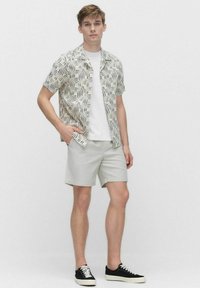 Korte mouwen shirt in crème met zwart golvend patroon, gecombineerd met lichtgrijze shorts en zwarte sneakers met witte zolen en veters.
