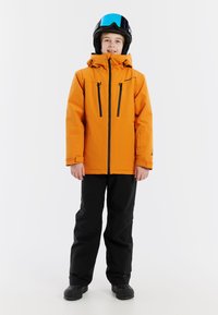 Oranje skijack met een capuchon, twee met rits afgesloten borstzakken en zwarte broek. Het model draagt een zwarte helm met een blauwe vizier en stevige zwarte laarzen.