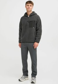 Fleece hoodie in donkergrijs met een rits en een zwarte borstzak; gecombineerd met grijze sweatpants en grijze sneakers.