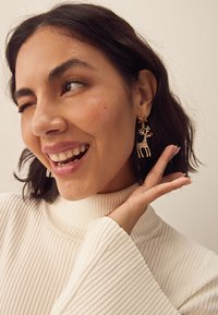 Gold-tone earrings shaped like a reindeer, adorned with red and white accents, paired with a white ribbed turtleneck sweater.