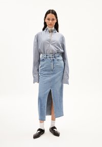 Lichtblauwe spijkerrok met een voorknoping, gecombineerd met een blauwe en witte gestreepte blouse met kraag en lange mouwen. Zwarte schoenen met gespen.