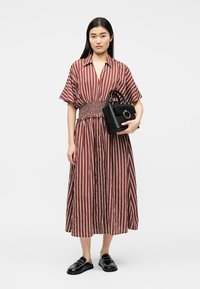 Γυναίκα που φοράει midi φόρεμα με καφέ και λευκές κάθετες ρίγες και ελαστική μέση, κρατώντας μια μαύρη τσάντα και φορώντας μαύρα παπούτσια χωρίς κορδόνια.