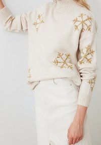 Beiger Pullover mit hohem Kragen, mit goldenem Schneeflocken-Muster auf den Ärmeln. Rippbündchen, weicher Stoff, kombiniert mit einem weißen Rock.